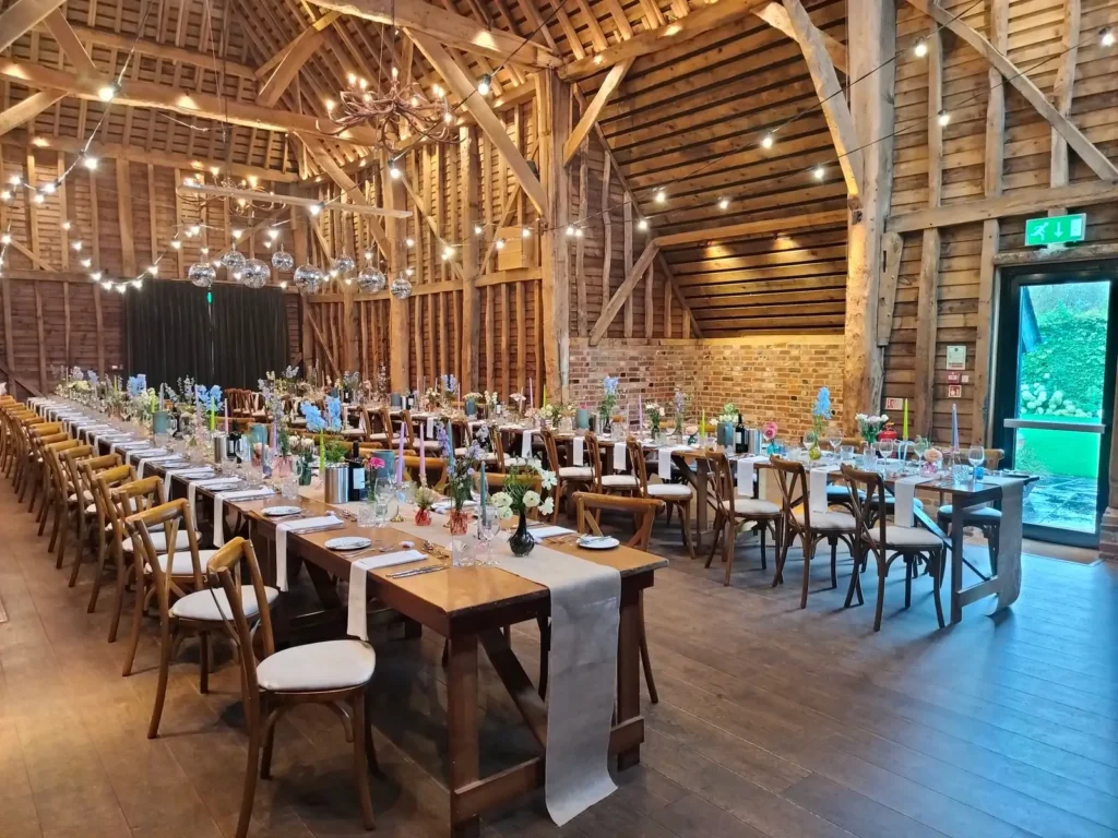Long wedding tables with flowers and festoon lights inside a timber barn